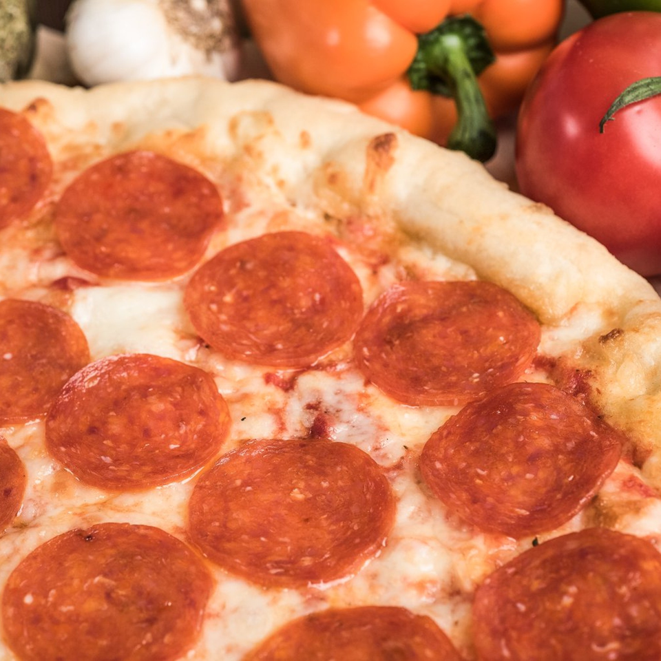 Close-up of a pepperoni pizza with melted cheese and a golden crust, with fresh vegetables in the background.